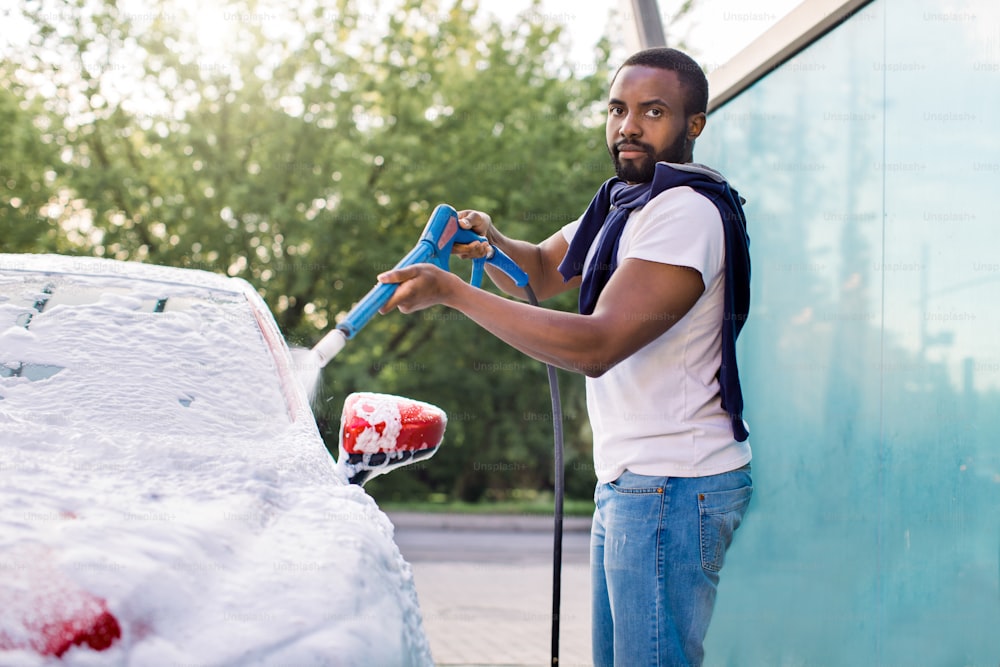 高圧ジェットを使用した石鹸泡による車の屋外清掃。ハンサムなひげを生やしたアフリカの若い男は、屋外のセルフウォッシュサービスで泡で高圧下で赤い車を洗っています。