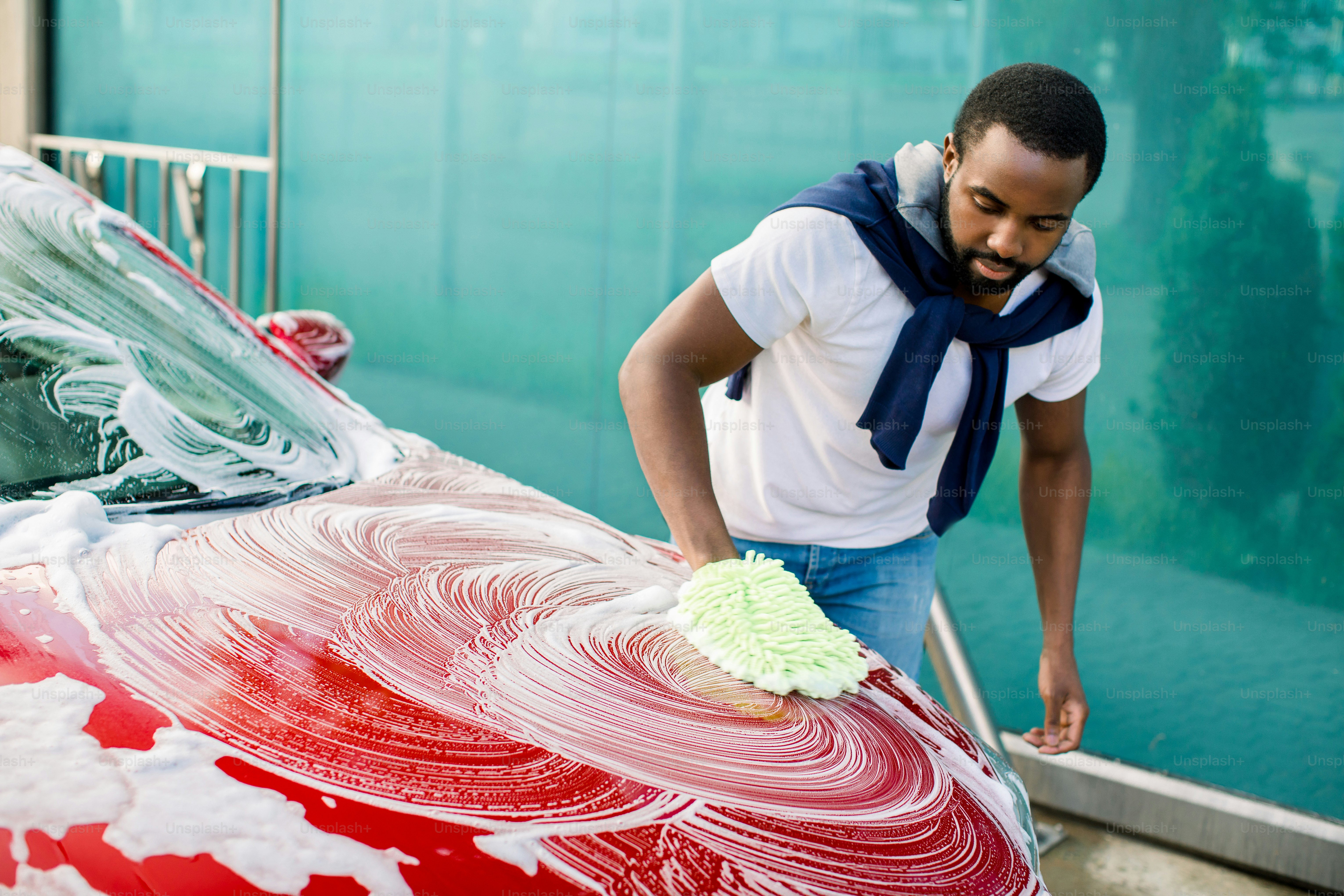 Horizontal view of car cleaning at self wash service outdoors. Handsome African man in casual wear washing red car with sponge and foam.