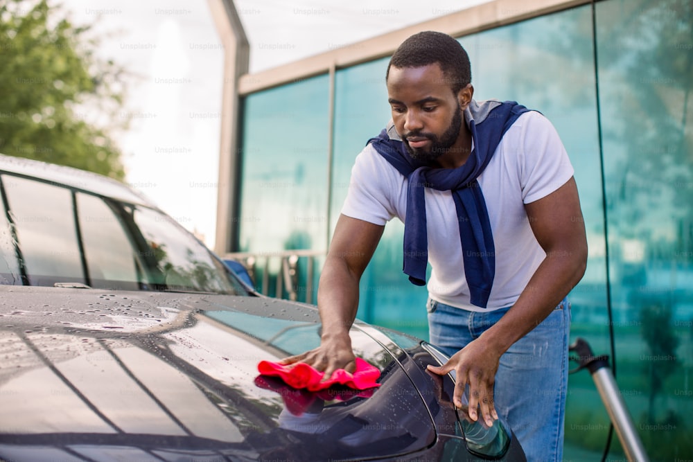 Scatto orizzontale di un giovane afroamericano che pulisce il cofano della sua moderna auto elettrica di lusso all'aperto con un panno in microfibra rossa, presso l'autolavaggio self-service.