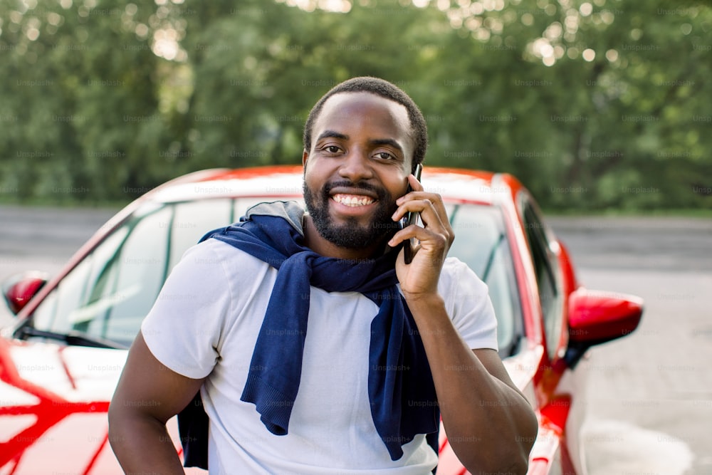 Uomo afroamericano bello positivo in piedi davanti alla sua lussuosa auto rossa e guardando la telecamera con un sorriso felice, mentre ha una conversazione telefonica all'aperto. Primo piano orizzontale.
