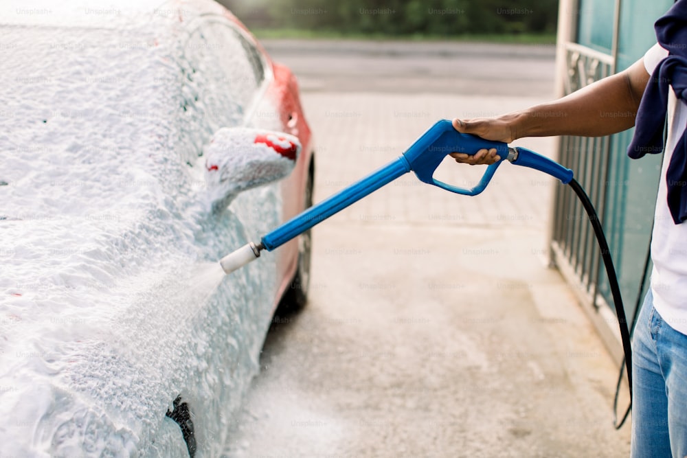 Car wash outdoors concept. Red car in foam. Car getting a wash with soap. Hand of African man with high pressure jet spraying the soap on red car at service station.