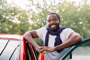 Atraente jovem africano sorridente em trajes casuais, posando para a câmera perto de seu novo carro vermelho de luxo ao ar livre na rua. Feliz proprietário do carro com seu carro vermelho.
