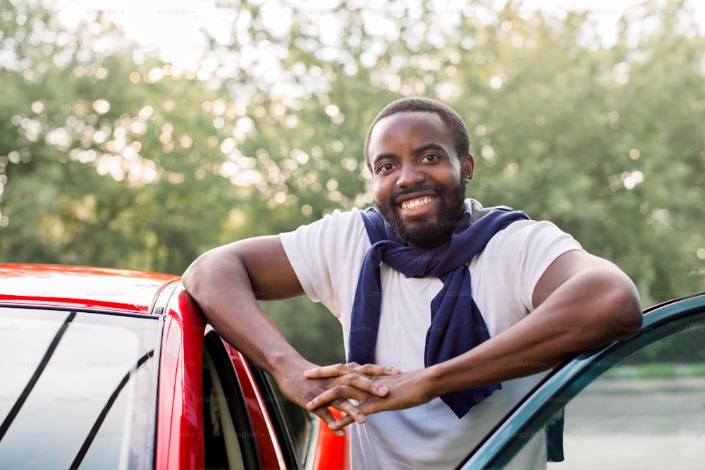 Atractivo joven africano sonriente en ropa casual, posando a la cámara cerca de su nuevo coche rojo de lujo al aire libre en la calle. Feliz propietario de un coche con su coche rojo.