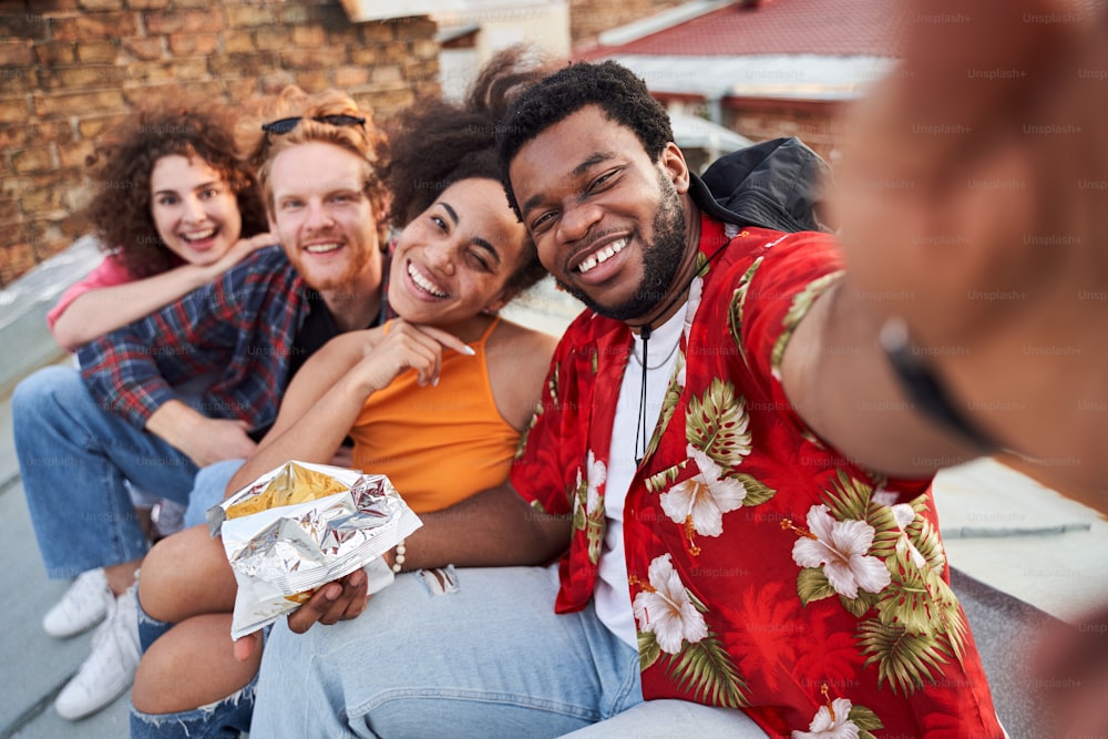 Porträt eines fröhlichen jungen Mannes, der Selfies mit Freunden macht, während sie sich umarmen und Chips essen