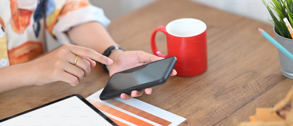 A cropped image of a woman is using a white blank screen smartphone at the wooden table.