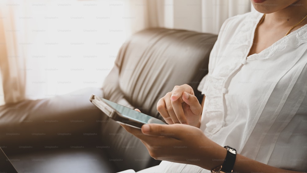 A cropped image of a woman is using a white blank screen smartphone at the black sofa.