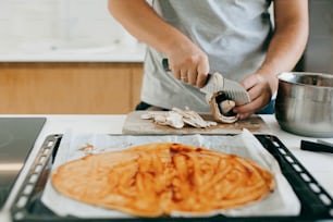 Person cutting mushrooms on background of round dough with ketchup for pizza on modern white kitchen. Process of making home pizza, ingredients close up. Home cooking concept