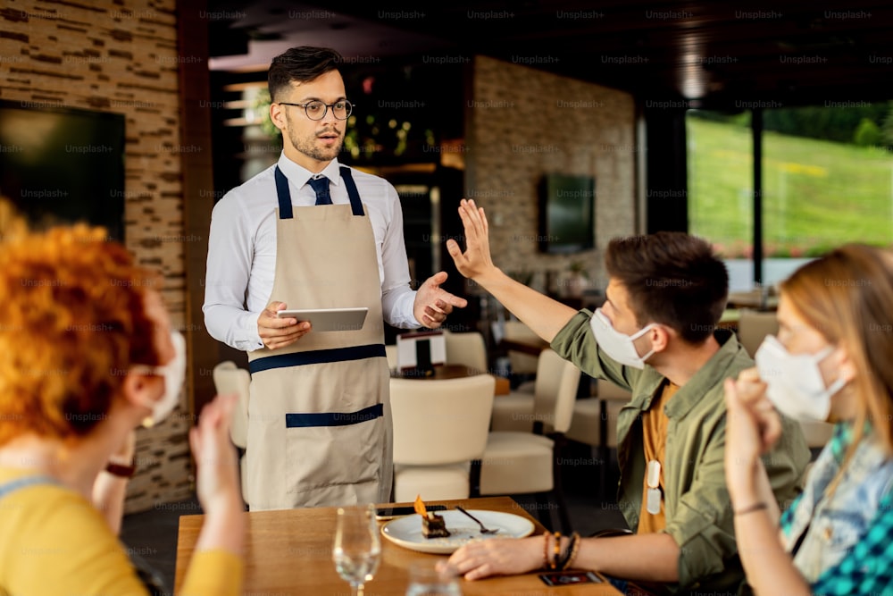 Grupo de clientes haciendo un gesto con una señal de alto a un camarero que no lleva mascarilla protectora en una cafetería durante la epidemia de coronavirus. La atención se centra en el camarero.