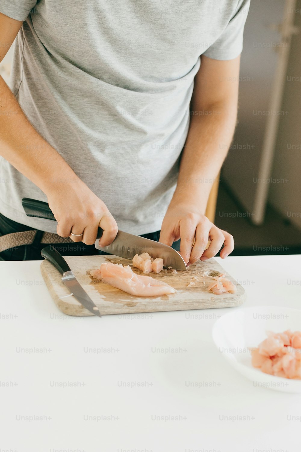 Persona cortando filete de pollo con cuchillo sobre tabla de madera en cocina blanca moderna. Proceso de elaboración de pizza casera, con las manos en alto. Concepto de cocina casera