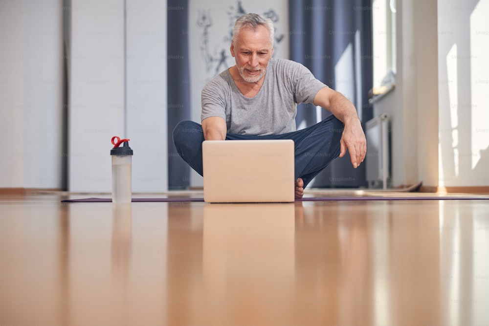 Hombre maduro sonriente y concentrado sentado con las piernas cruzadas en la esterilla de yoga frente a su computadora