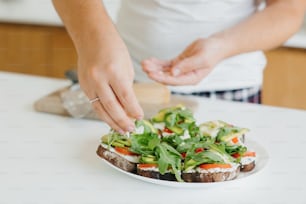 Sandwich made of whole grain bread, avocado, tomato, arugula and cheese. Home cooking concept. Hands putting parmesan on homemade toasts with avocado on modern white kitchen.