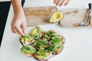 Hand putting arugula on sandwich made of whole grain bread, avocado, tomato, and cheese. Process of making toasts with avocado on modern white kitchen. Home cooking concept