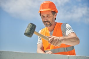 Waist-up portrait of a smiling pleased mature Caucasian mason using a hammer for laying bricks