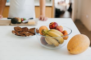 白いモダンなキッチンでアボカド、チェリートマト、パンでトーストする人の背景に果物が描かれたスタイリッシュなガラスのボウル。カウンタートップにバナナ、桃、アプリコット、メロン