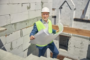 Vista frontal de um alegre engenheiro sênior do sexo masculino em um colete de segurança segurando os planos da casa