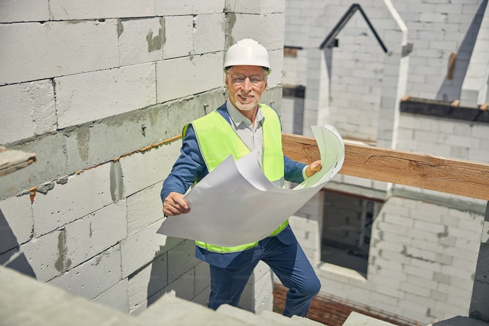 Front view of a joyous senior male engineer in a safety vest holding house plans