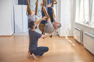 Professeur de yoga fort soutenant un homme aux cheveux gris suspendu dans un hamac en soie pendant l’entraînement