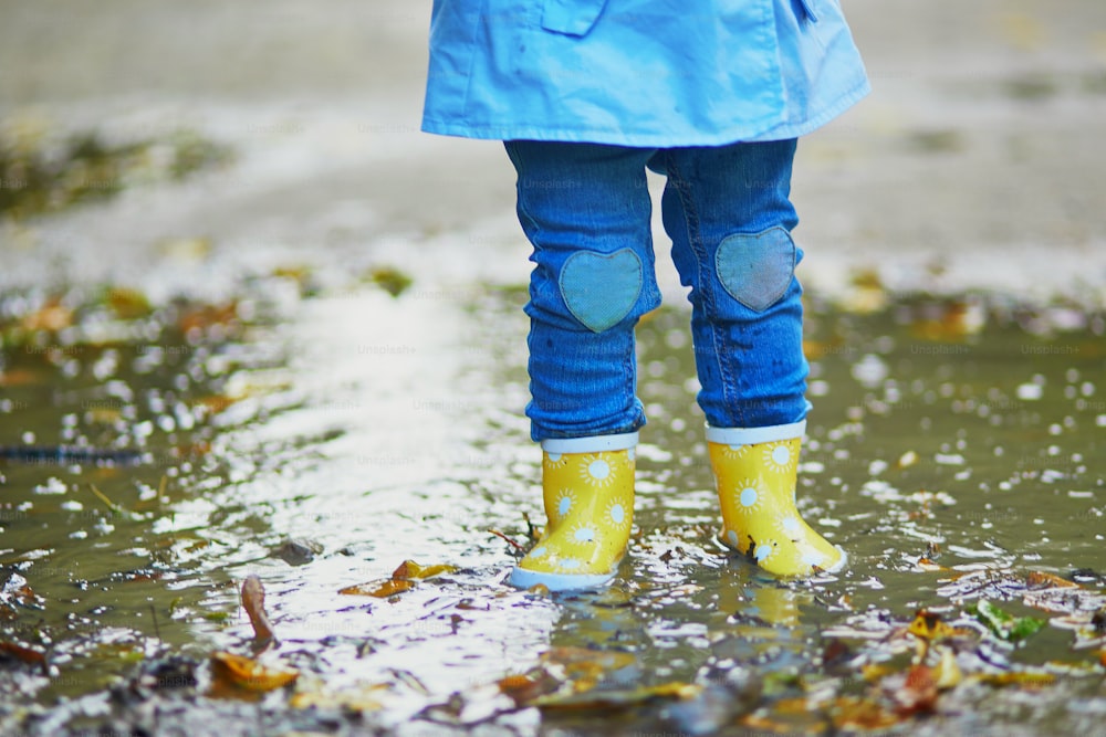 秋の日に黄色いレインブーツを履いて水たまりに飛び込む子供。雨の日に公園で水と泥で楽しんでいる幼児の女の子。子供のための秋の野外アクティビティ