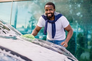 Lavaggio auto, concetto di servizio di autolavaggio auto all'aperto. Giovane africano sorridente in abbigliamento casual che pulisce la sua nuova auto moderna usando spugna e schiuma, guardando la macchina fotografica.