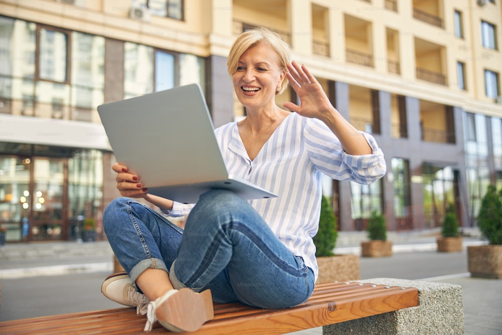 High-spirited attractive mature blonde lady sitting cross-legged with a computer on a wooden bench outside