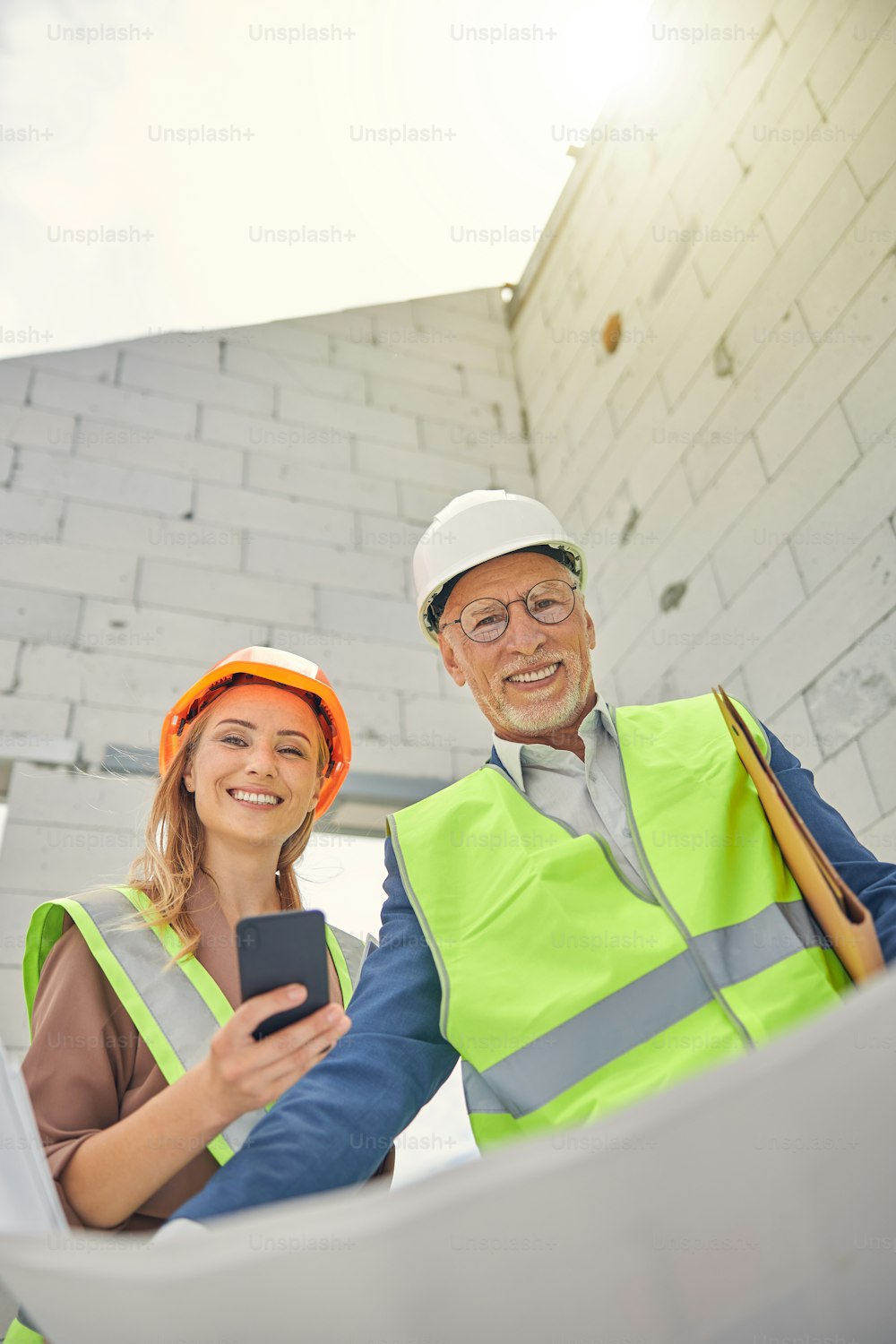 Contre-plongée d’une femme souriante avec un smartphone debout à côté d’un ingénieur civil