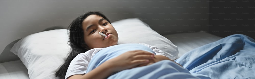 A little sick girl in a blanket is lying on the bed and measuring the temperature by her self.