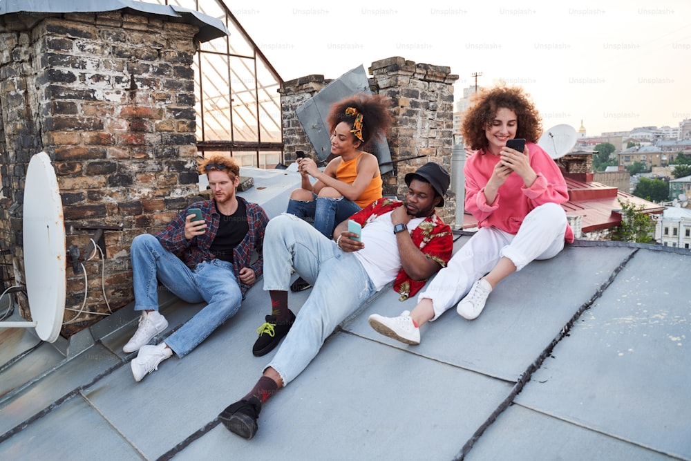 Glückliche vier Männer und Frauen entspannen sich auf dem Stadtdach und nutzen Smartphones für Messaging