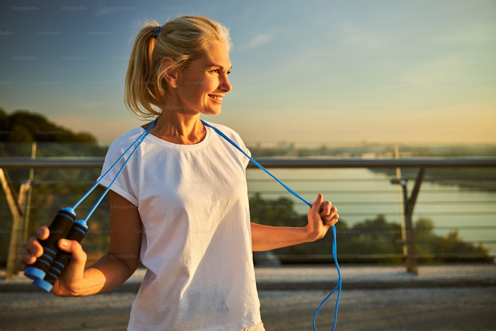 Charming lady in white shirt holding jump rope and smiling while spending time outdoors