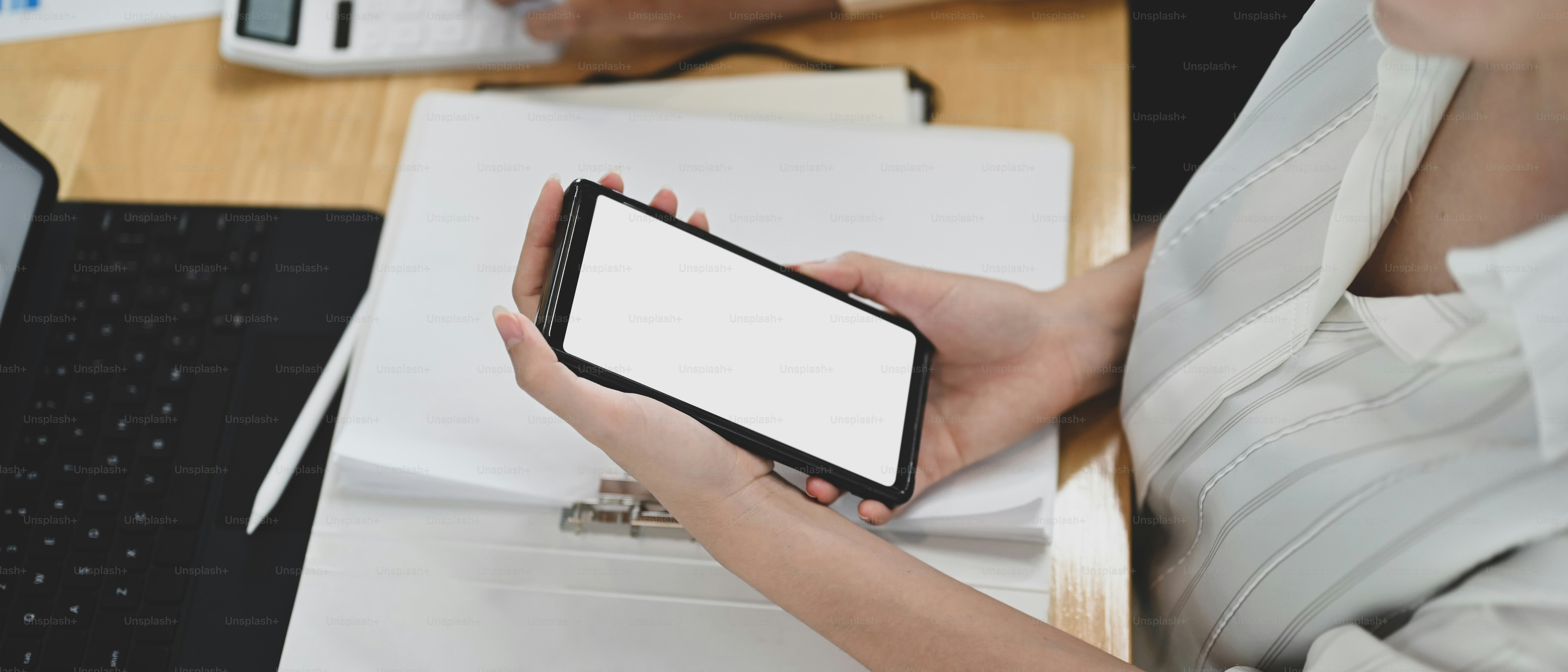 A woman is using a white blank screen mobile phone while sitting in front of a computer laptop at the wooden table.