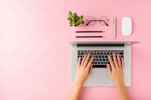 Female hands working on modern laptop on pink background with accessories. Office desktop. Flat lay