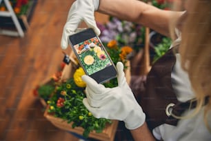 작업 장갑을 낀 금발의 백인 여성이 스마트폰으로 꽃을 촬영하는 모습