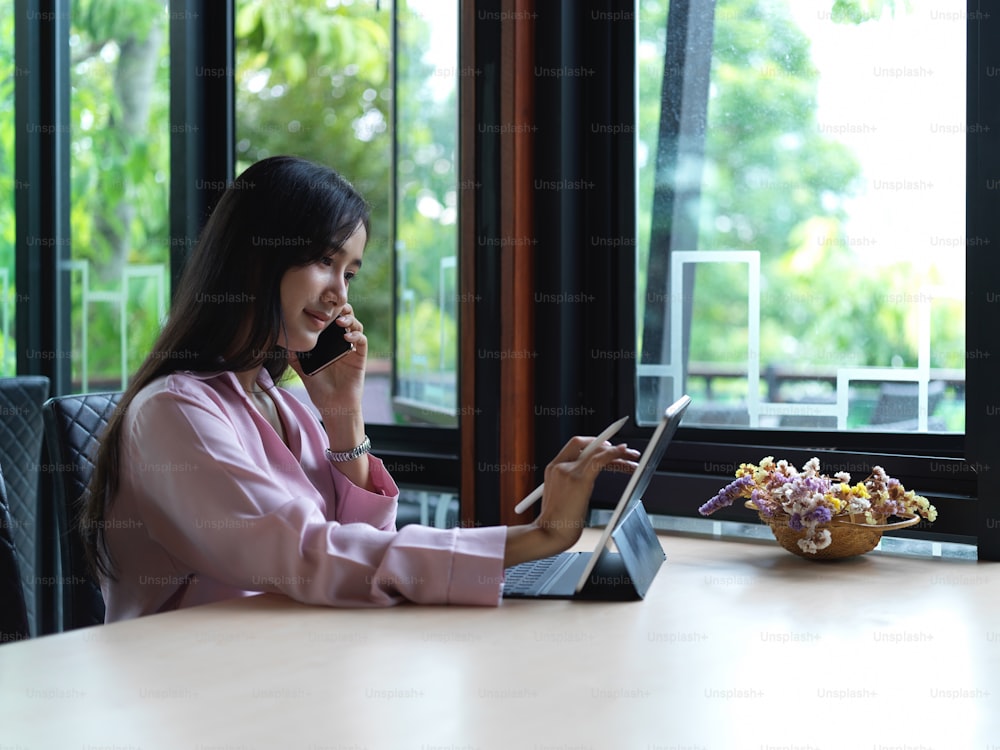 Side view of businesswoman talking on phone while working with digital tablet on table in cafeteria