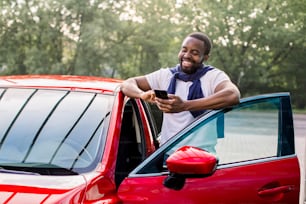 Outdoor lifestyle portrait of satisfied laughing young African American man in t-shirt using mobile phone while standing at his modern red car. Happy man using car apps.