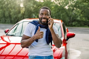 Joyful satisfied African young man in casual wear, standing outdoors in front of his red car, talking on mobile phone and showing his thumb up to camera. Good auto service concept, copy space.