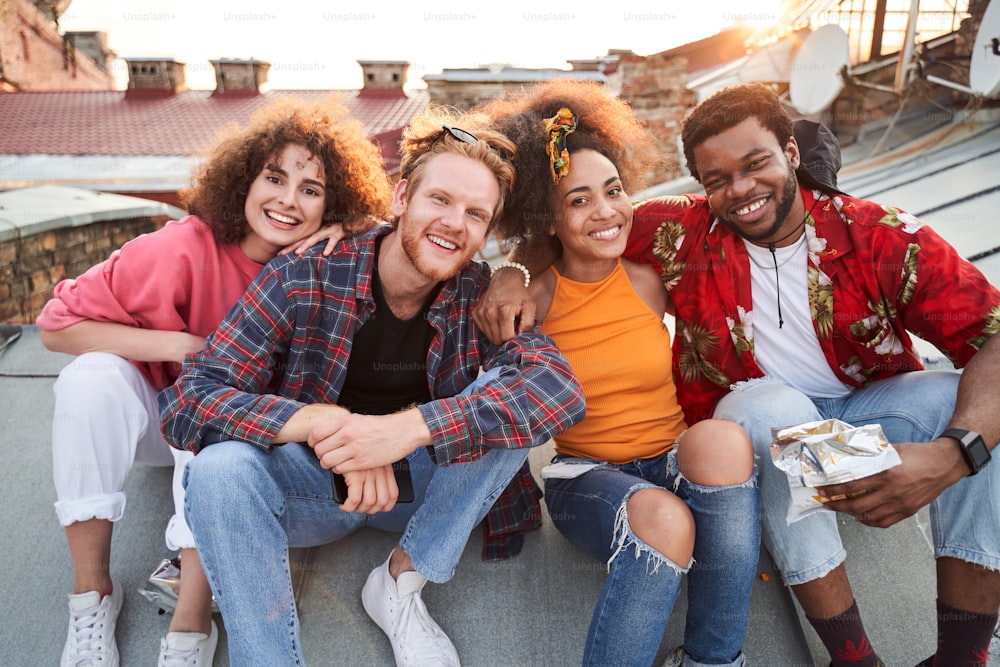 Full length portrait of four happy young people hugging and relaxing while eating chips outdoors
