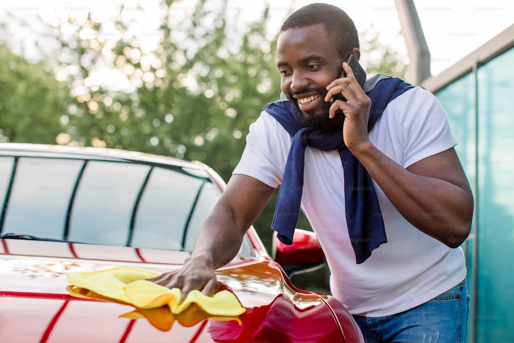 실외, 셀프 서비스 스테이션에서 세차. 젊은 바쁜 아프리카 남자가 노란색 극세사 천으로 빨간 차를 닦는 동안 전화를 걸고 있다