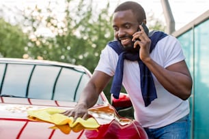 Lavaggio auto all'aperto, stazione self service. Giovane uomo africano occupato che parla al telefono mentre lucida la sua auto rossa con un panno in microfibra giallo