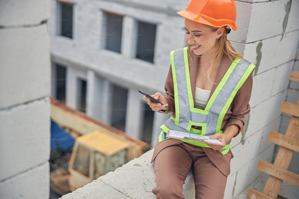 Front view of a cute merry young blonde female worker leaning against the brick wall