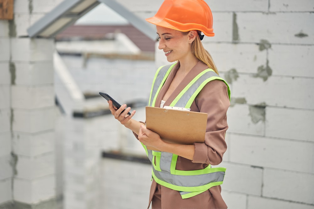 Side view of a smiling young female constructor reading a text message on her smartphone