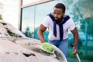 Giovane uomo afroamericano attraente che lava la sua moderna auto elettrica di lusso in una stazione di autolavaggio self-service all'aperto con schiuma detergente e guanto di spugna verde.