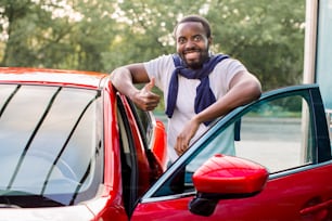 Joven hombre afroamericano alegre y sonriente con camiseta blanca y suéter alrededor del cuello, de pie cerca de su nuevo automóvil rojo de lujo al aire libre y mostrando el pulgar hacia arriba.