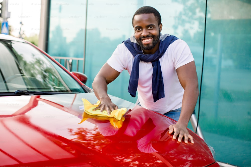 Ritratto di un giovane sorridente dalla pelle scura che pulisce il cofano rosso della sua auto con un panno in microfibra gialla all'aperto, alla stazione di autolavaggio di auto.