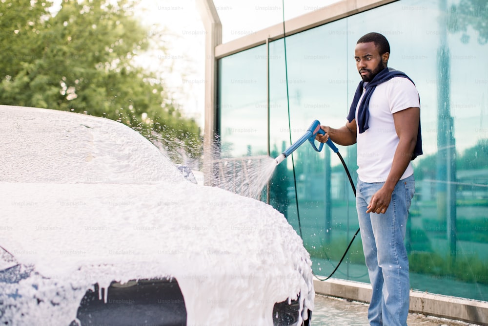 Outdoor cleaning of the car with soap foam using high pressure jet. Handsome bearded African young man washing his modern car under high pressure with foam in self wash service outdoors.