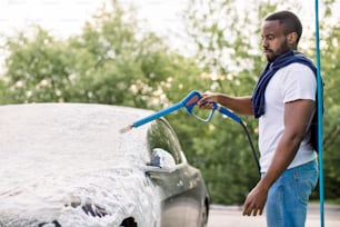 Joven negro limpiando su automóvil eléctrico de lujo gris con espuma en aerosol afuera en la estación de lavado de autos. Chico africano casual lavando su coche con rociador de alta presión y espuma.
