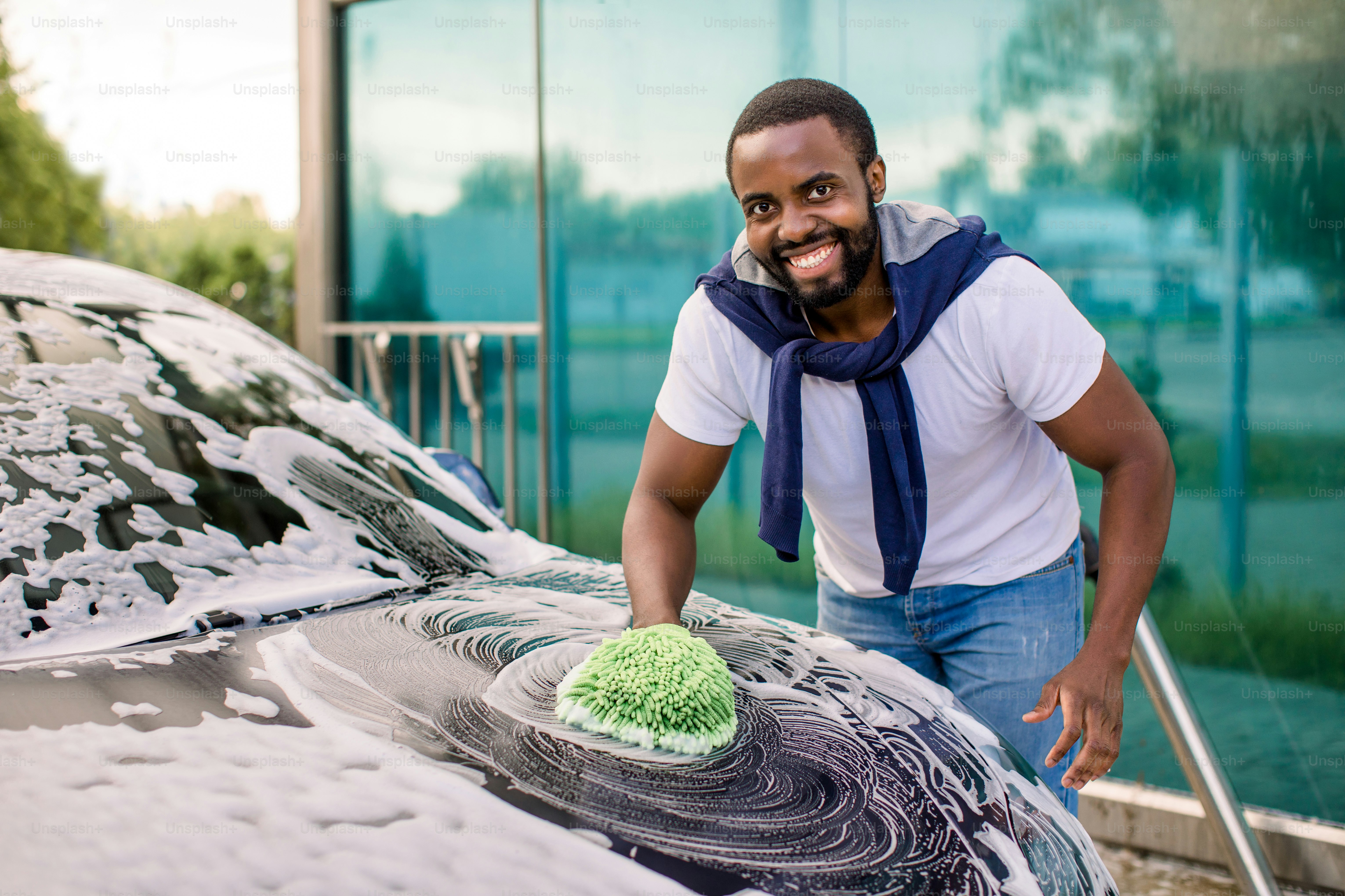 Horizontal view of car cleaning at self wash service outdoors. Handsome African man in casual wear looking at camera with happy smile while washing the car with sponge and foam