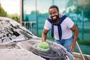 Vista orizzontale della pulizia dell'auto presso il servizio di autolavaggio all'aperto. Bello uomo africano in abbigliamento casual che guarda la macchina fotografica con un sorriso felice mentre lava l'auto con spugna e schiuma
