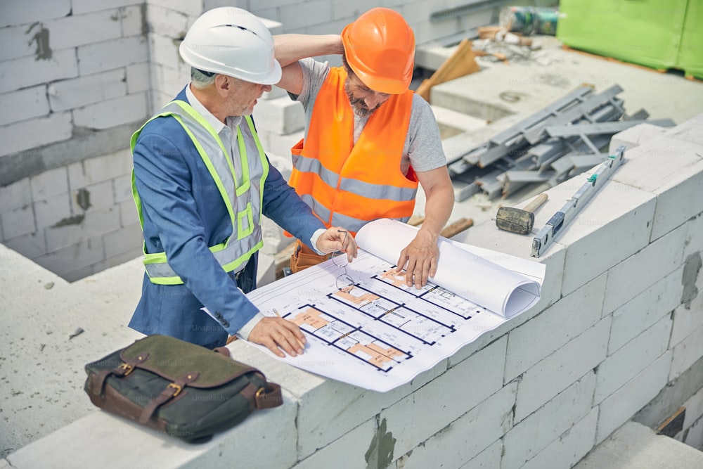 Grey-haired foreman with eyeglasses and a pencil in his hands talking to a perplexed builder