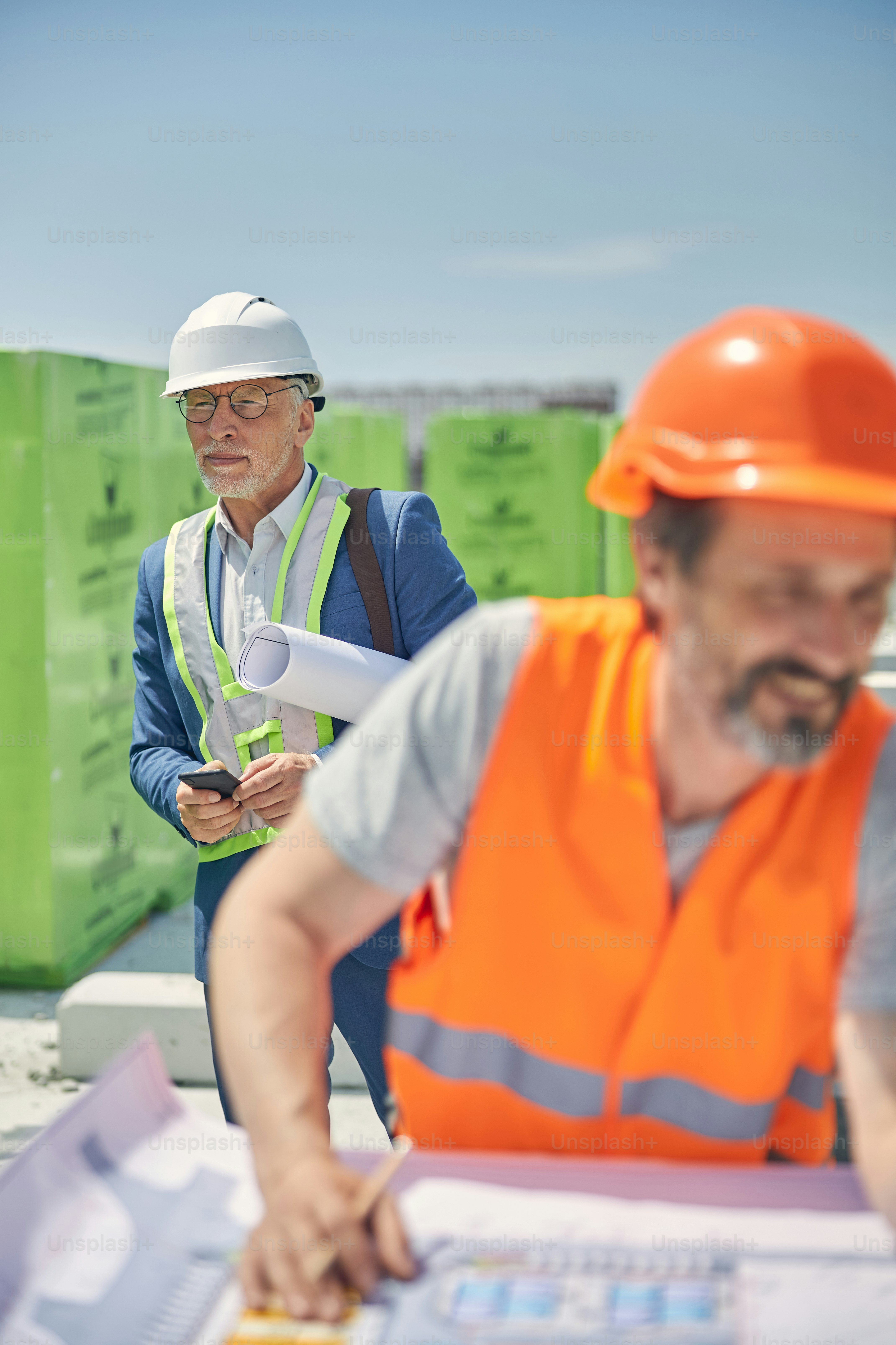 Ernster Mann mit einer Papierrolle unter dem Arm steht hinter einem lächelnden Ingenieur