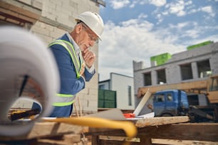 Serious calm Caucasian male engineer pondering over his house plans lying on a wooden table