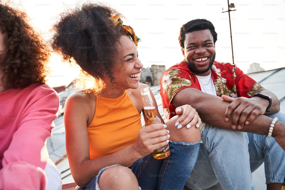 Merry male and female are sitting with friends and relaxing with bottles of beer at sunset